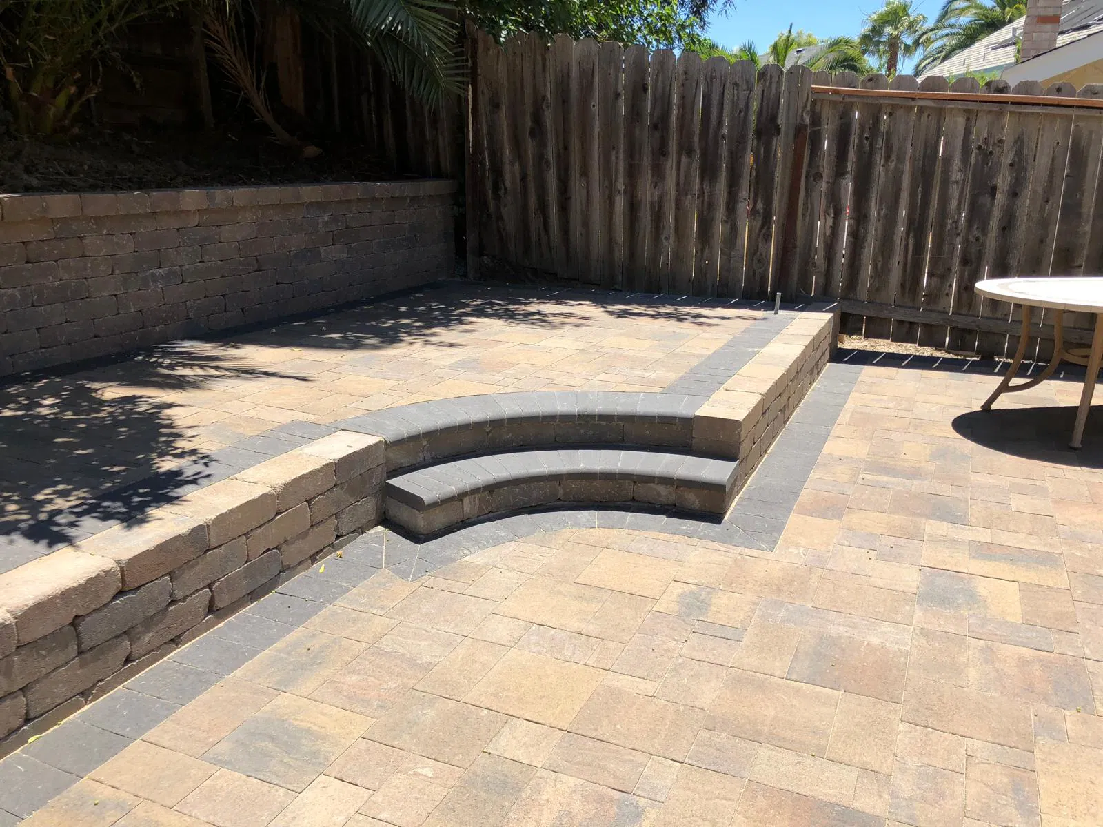Sunlit patio with paver stone bricks, curved steps, a raised wall, and a wooden fence in the background—expertly crafted by a leading paving installer in the Bay Area.