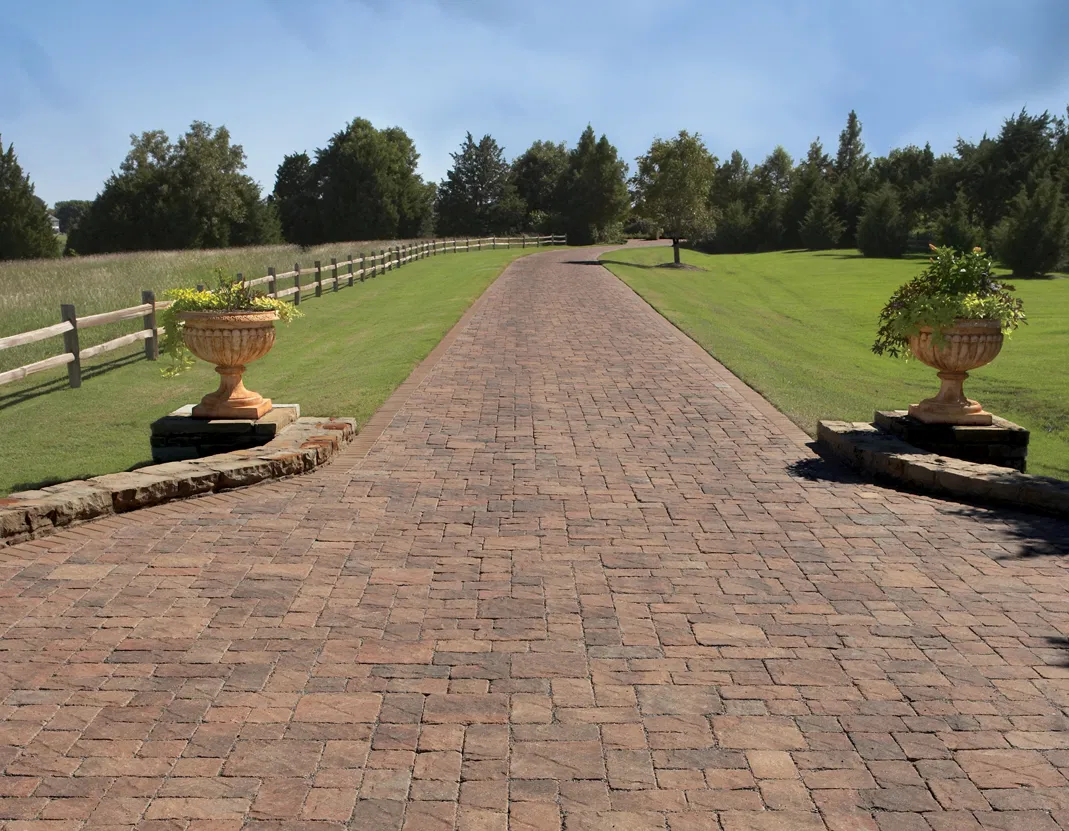 A long brick driveway flanked by grassy lawns, trees, and two large stone planters with greenery showcases the work of an expert paving installer specializing in paver stone design.