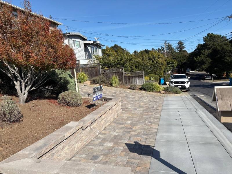 A paved sidewalk and driveway with interlocking paving, landscaping, a tree, a house, and a white truck parked on the street.