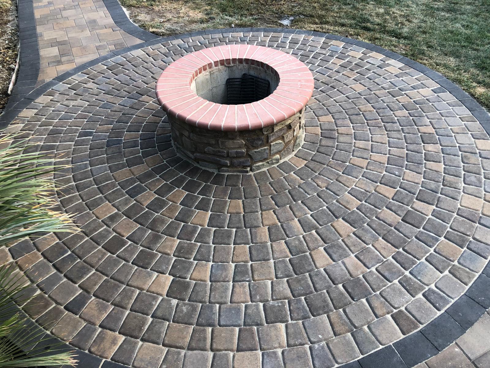 Circular brick fire pit surrounded by concentric interlocking paving stones with a curved pathway nearby.