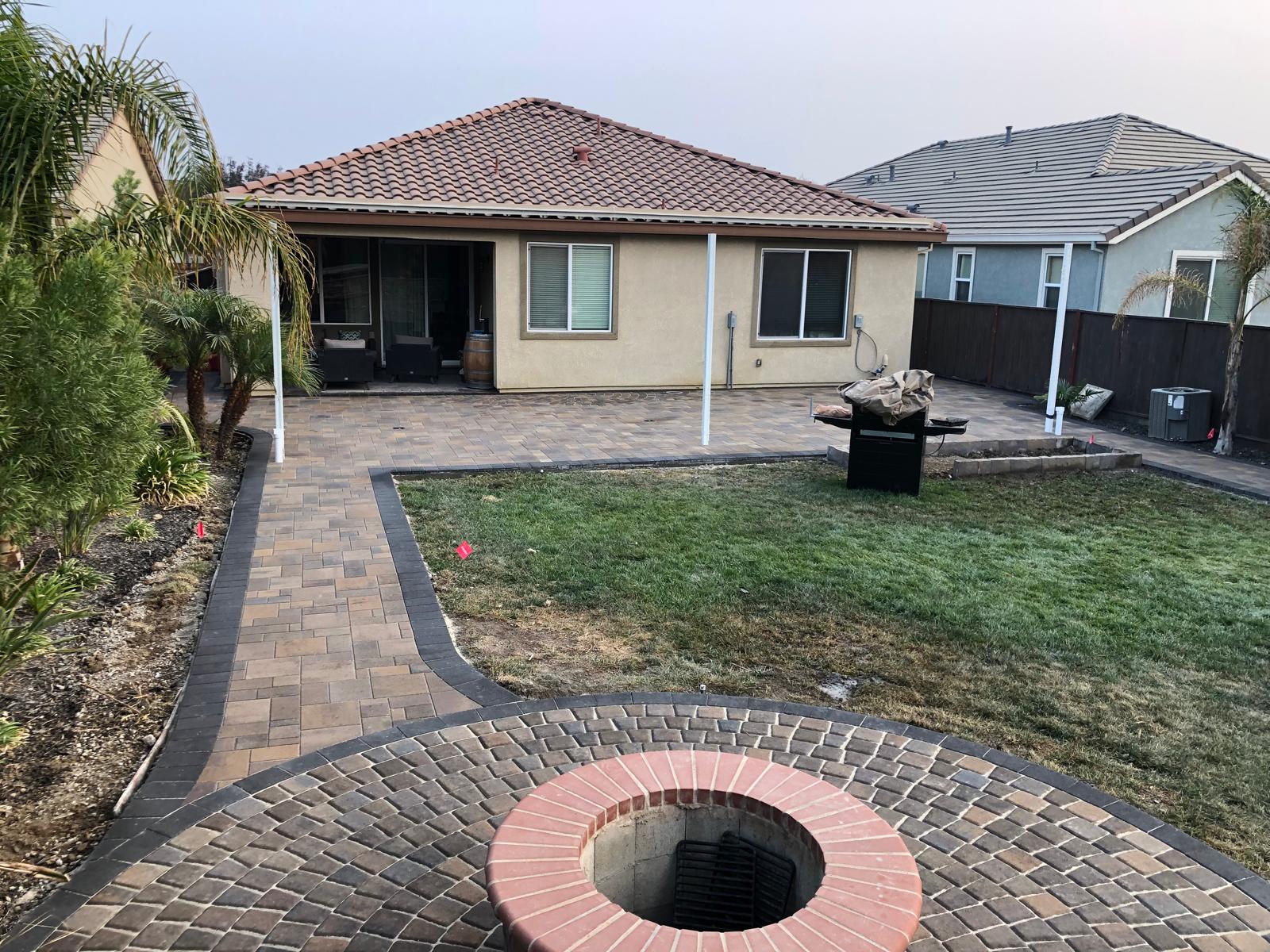 Backyard with brick patio, lawn, fire pit, palm trees, and a beige house with a red tile roof features attractive paver stone accents.