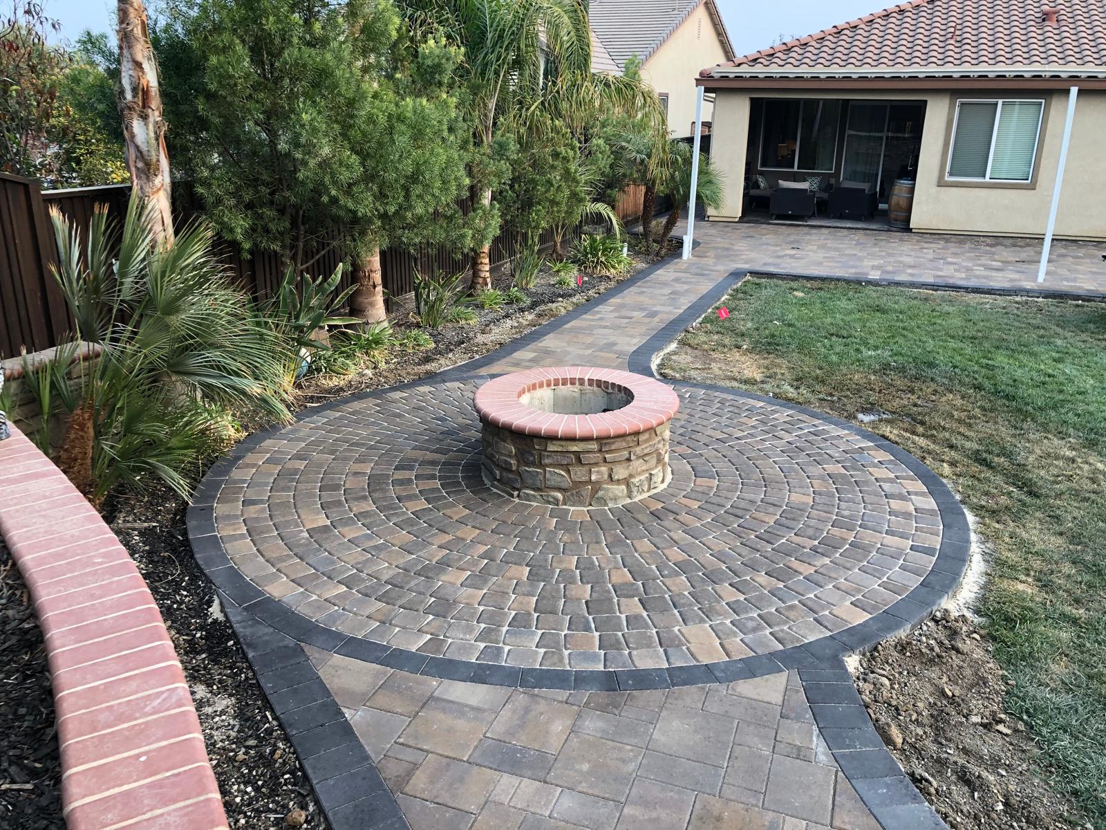 Circular stone fire pit with interlocking paving and brick patio set in a landscaped backyard near a house in the Bay Area.