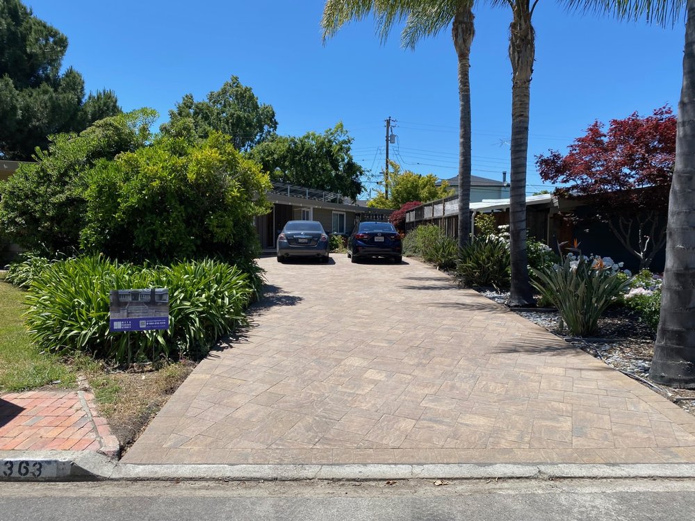 A bay area driveway with two parked cars, surrounded by plants and trees, leads to a house on a sunny day, showcasing expert work by a local paving installer.