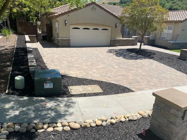 Single-story house with a large driveway installed by a skilled paving contractor, mulch landscaping, and a green utility box by the sidewalk.