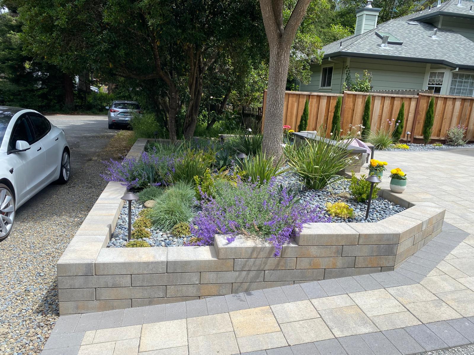 Landscaped garden bed with purple flowers and green plants, bordered by a light brick wall near a driveway, designed by an expert paver contractor.