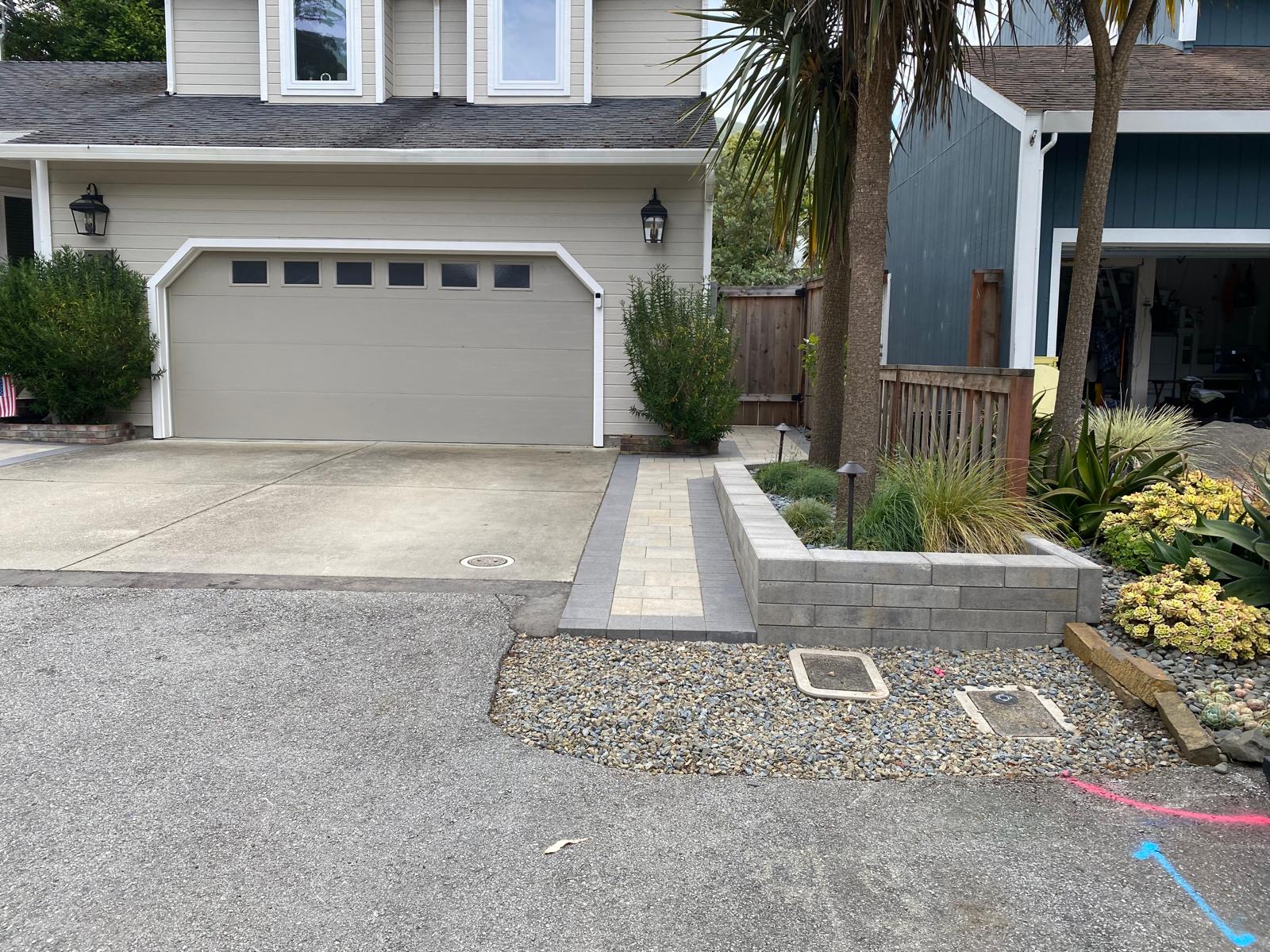 Modern gray driveway with a raised paver stone walkway, bordered by plants and a palm tree in front of a Bay Area house.