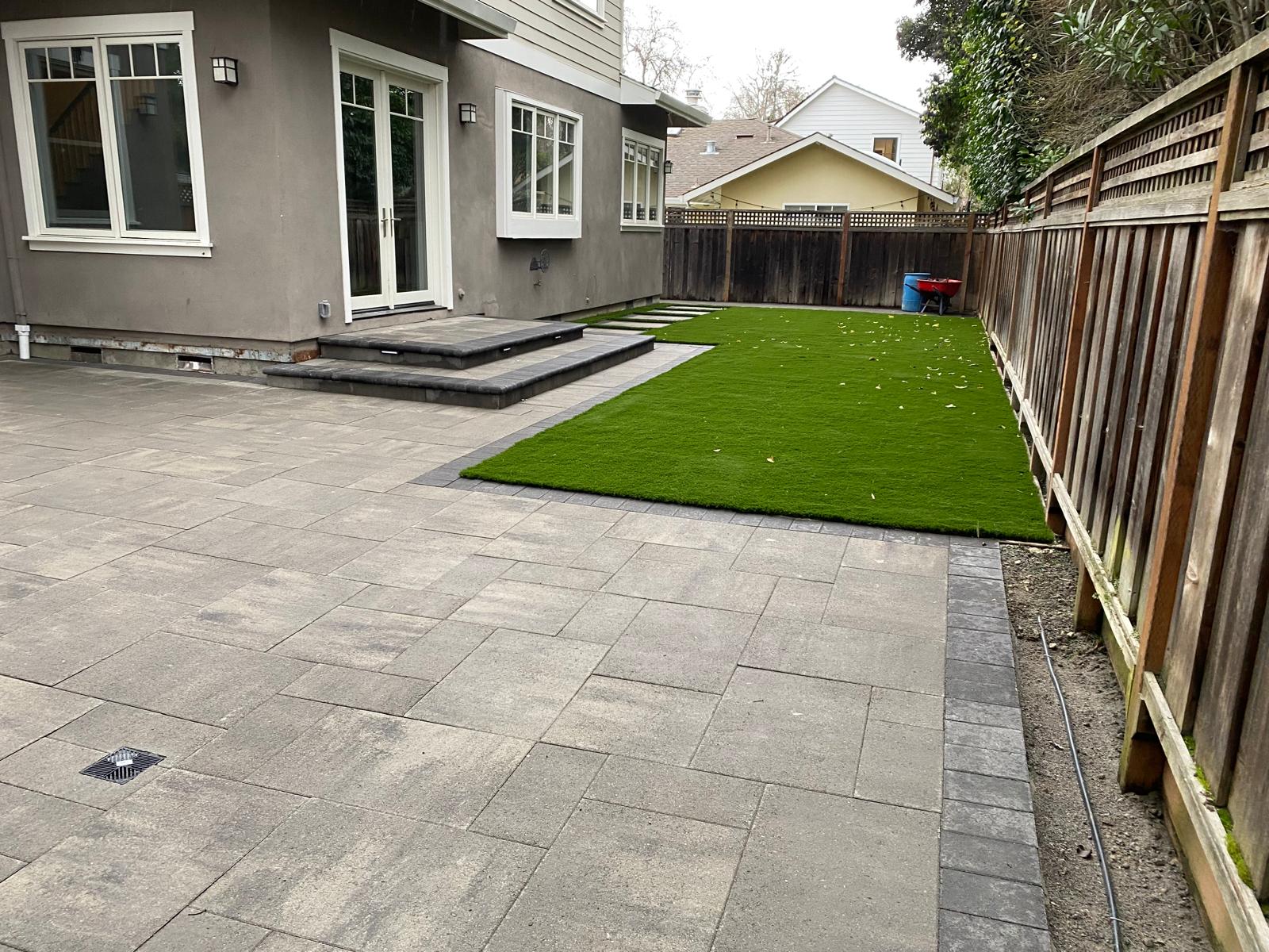 Backyard with a paver stone patio, artificial grass, wooden fence, and steps leading to glass doors of a bay area house.