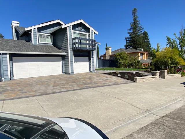 Two suburban houses with driveways featuring interlocking paving, surrounded by trees and a clear blue sky in a Bay Area residential neighborhood.