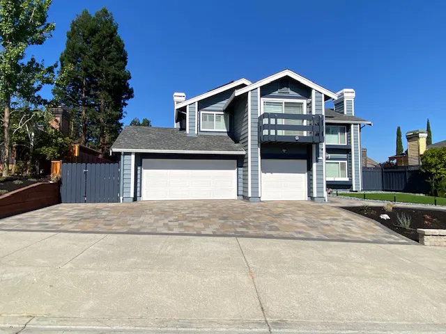 Two-story blue house with a three-car garage, wide driveway featuring interlocking paving by a skilled paver contractor, and clear blue sky in the background.