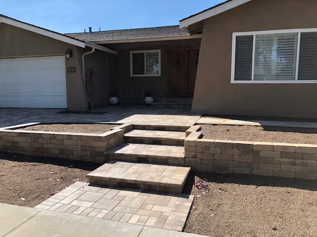 Front view of a house featuring steps, a stone walkway with paver stone accents, raised beds, and bare soil in the yard.