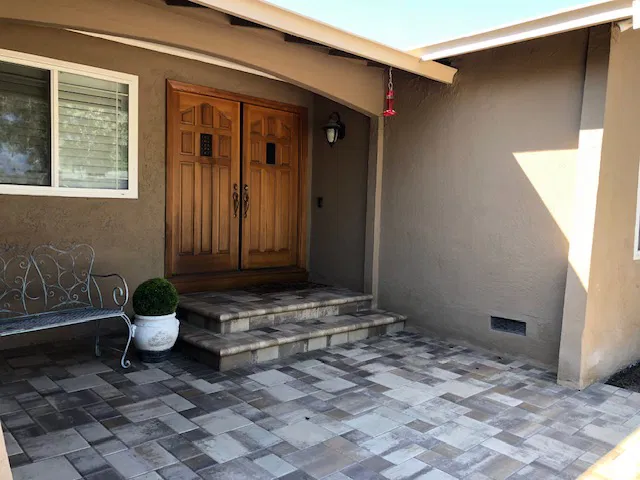 Front porch with tiled steps, interlocking paving, double wooden doors, a window, a bench, and a potted plant.