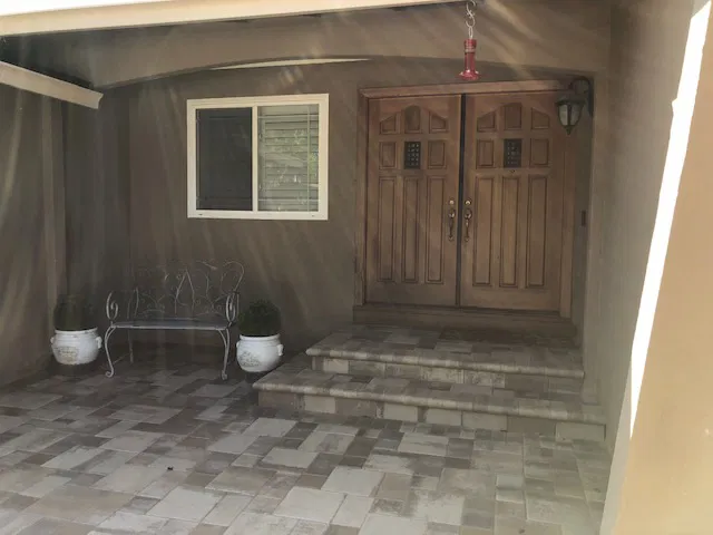 Wooden double doors, interlocking paving on the porch, metal bench, two potted plants, and a window beside the entrance by a trusted Bay Area paver contractor.