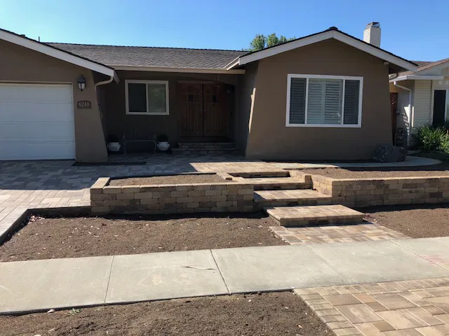 Single-story house with tan walls, a brick walkway featuring interlocking paving steps, and a bare soil front yard without grass or plants.