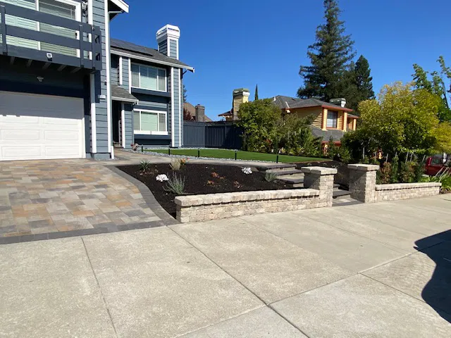 Modern Bay Area house with gray siding, stone driveway by a skilled paver contractor, small retaining wall, and landscaped front yard on a sunny day.