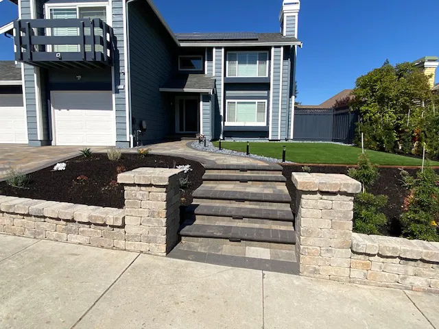 Stone steps with brick pillars and interlocking paving lead to a modern two-story house surrounded by a manicured lawn and garden beds.