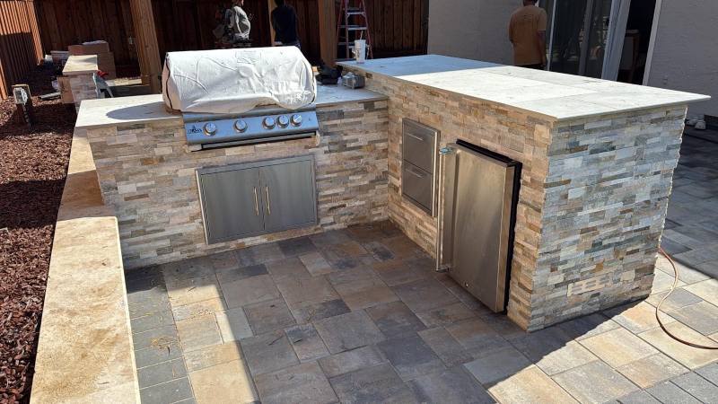 Outdoor kitchen with a built-in grill, fridge, storage, and stone finish on an interlocking paving patio by a trusted paver contractor.