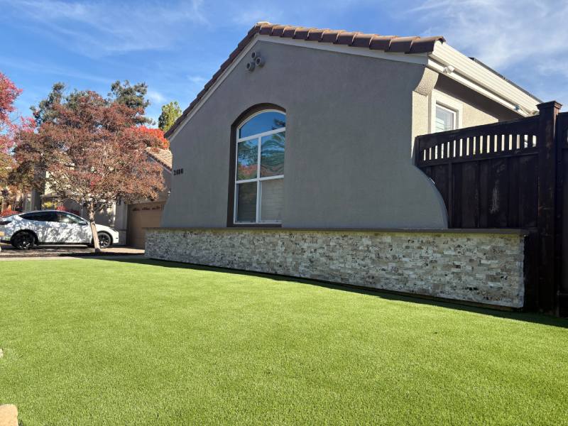 Single-story house with arched window, stone accent wall, artificial grass lawn, and expertly installed paving by a Bay Area paver contractor under a blue sky.