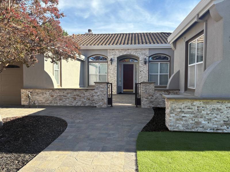 Single-story house with stone accents, arched windows, and a tree by the walkway featuring elegant paver stone detailing under a partly cloudy sky.
